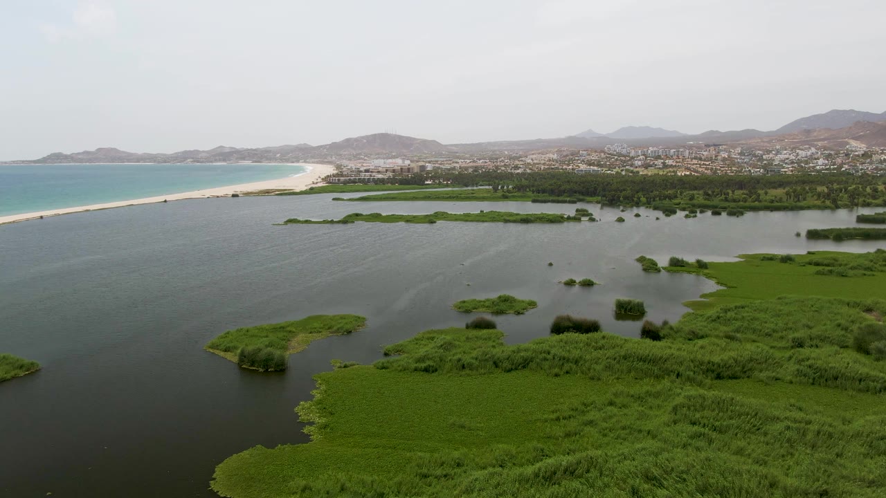 Mid-clip aerial over Puerto Los Cabos Beach shows the lagoon opening wider across the frame, with marsh vegetation at the bottom, a long sandy beach at left, turquoise ocean, distant settlement, hills, and no visible people.