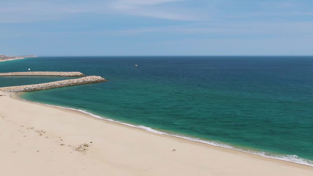 Elevated view of Puerto Los Cabos Beach with empty sand below, darker turquoise ocean beyond the shore, the rocky breakwater extending from the left, and no visible people.
