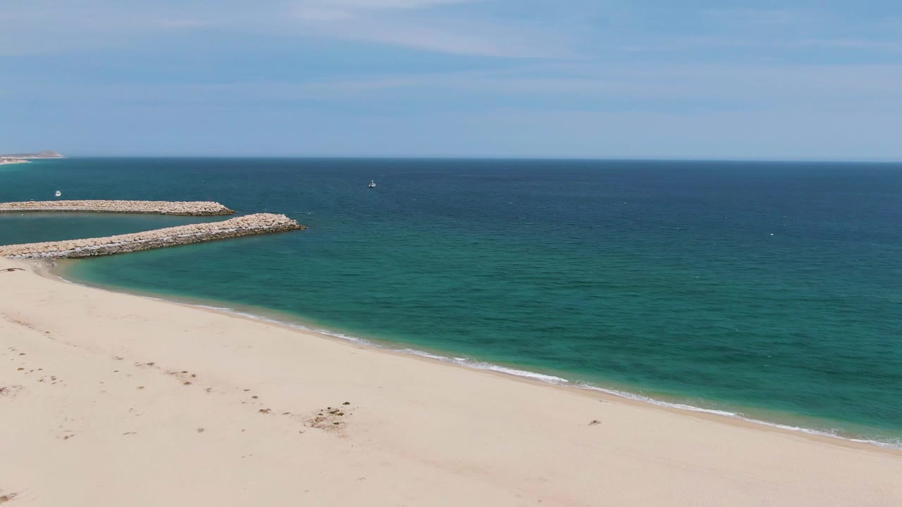 Puerto Los Cabos Beach in Baja California Sur with pale sand in the foreground, turquoise shoreline water, a rocky breakwater at left, and no visible people.