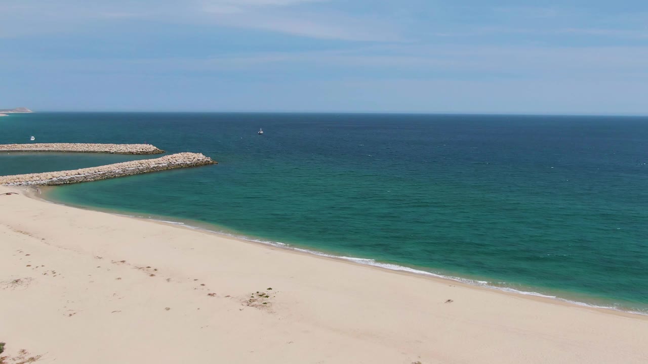Puerto Los Cabos Beach aerial view with the rocky jetty extending into the ocean, a smooth sandy foreground, gentle shore foam, and no visible people.