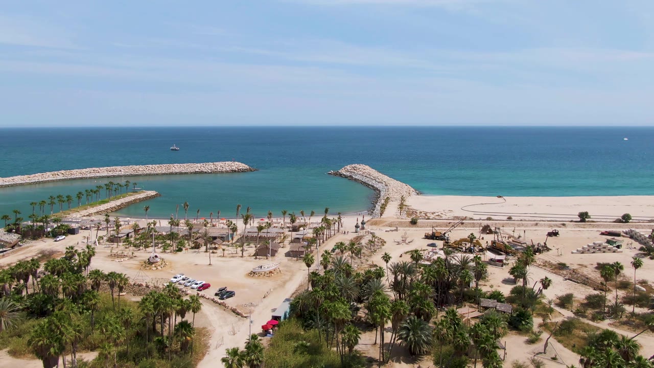 Wide aerial view at Puerto Los Cabos Beach with the red vehicle farther along the sandy road, parked cars near palms, breakwaters in the water, and no visible people.
