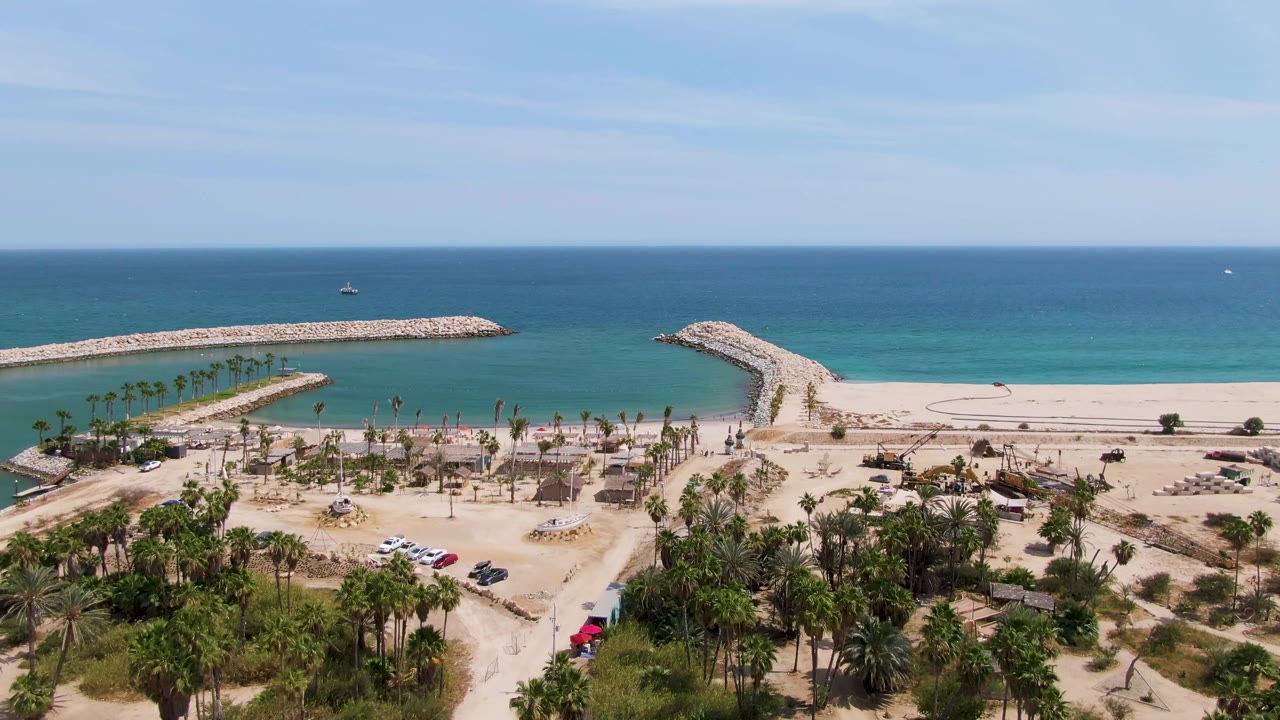 Aerial still of Puerto Los Cabos Beach with the sandy road and palm grove below, the red vehicle near the lower right road area, rocky breakwaters offshore, and no visible people.