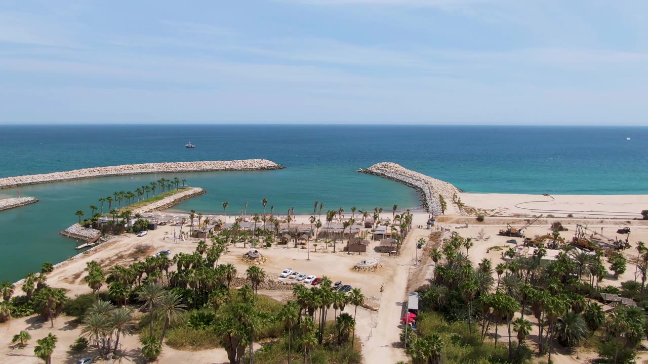 Aerial frame at Puerto Los Cabos Beach shows turquoise water, two rocky breakwaters, sandy beach, palm trees, a road with parked vehicles, small boats offshore, and no visible people.