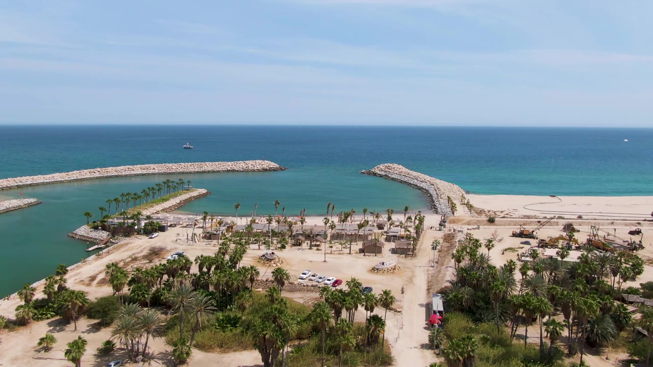At 0:01, the Puerto Los Cabos Beach aerial view holds on the narrow channel, with palms and parked vehicles below, rock breakwaters around the inlet, and no visible people.