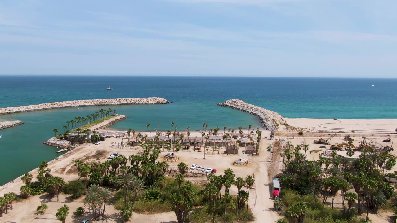 At 0:02, the drone perspective over Puerto Los Cabos Beach shows foreground palms and road, a sheltered turquoise channel, curved rock breakwaters, open ocean beyond, and no visible people.