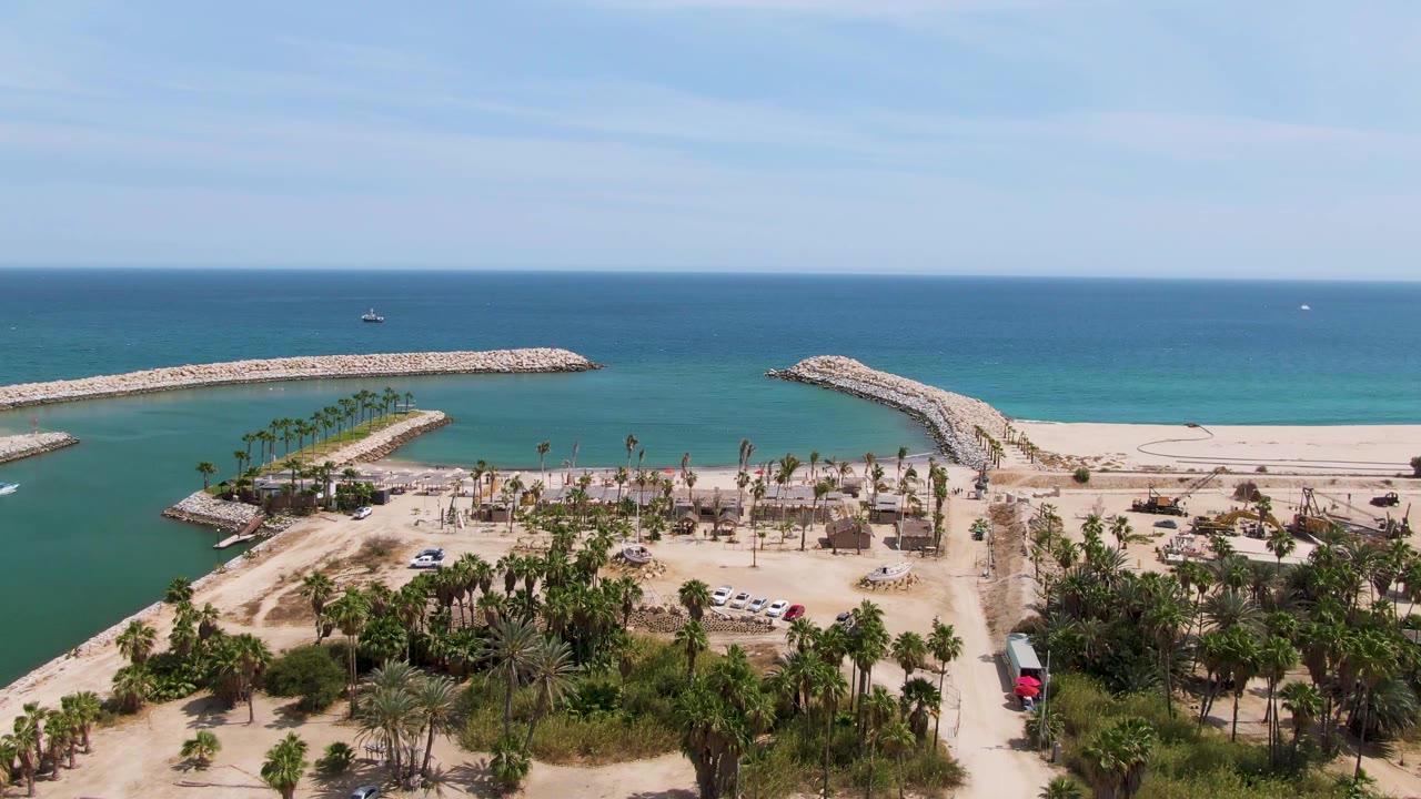Drone frame of Puerto Los Cabos Beach channel with breakwaters, palms, sand, and calm ocean.