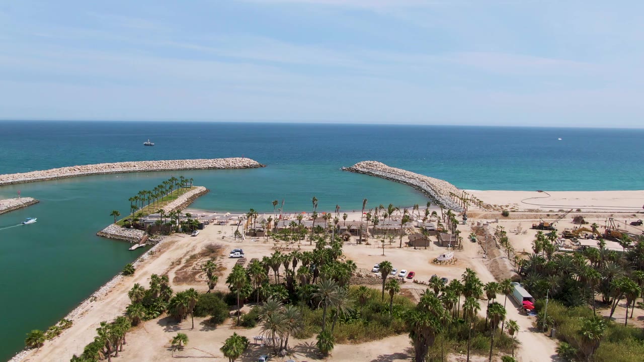 Aerial view of breakwaters, channel water, sandy beach, palms, and ocean horizon.
