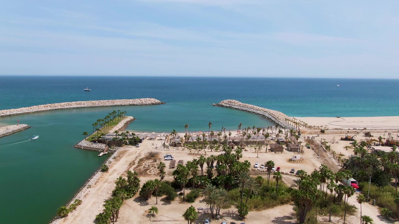 At 0:02, the Puerto Los Cabos Beach frame keeps the breakwater channel centered, with turquoise and green water around the sand, clustered palms, a few vehicles, and no visible people.