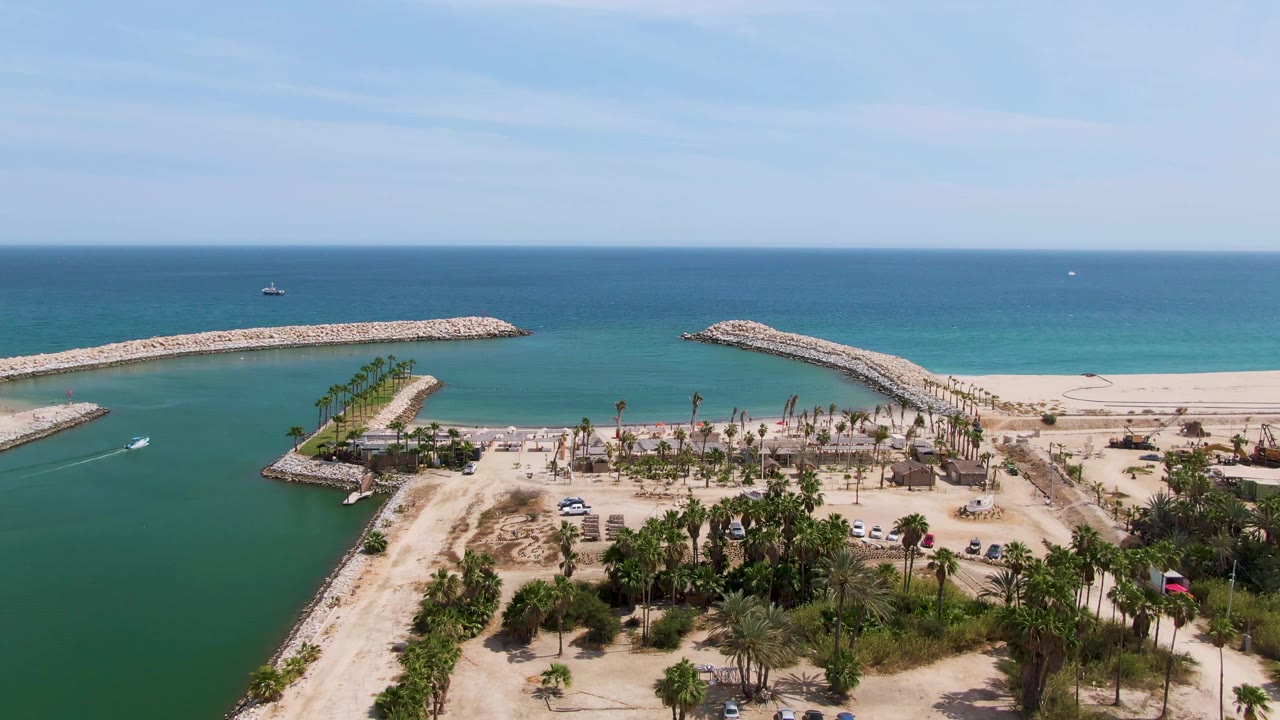 At 0:03, the Puerto Los Cabos Beach aerial frame shows more foreground parking and palms below the channel, with the breakwater arms leading toward the ocean and no visible people.