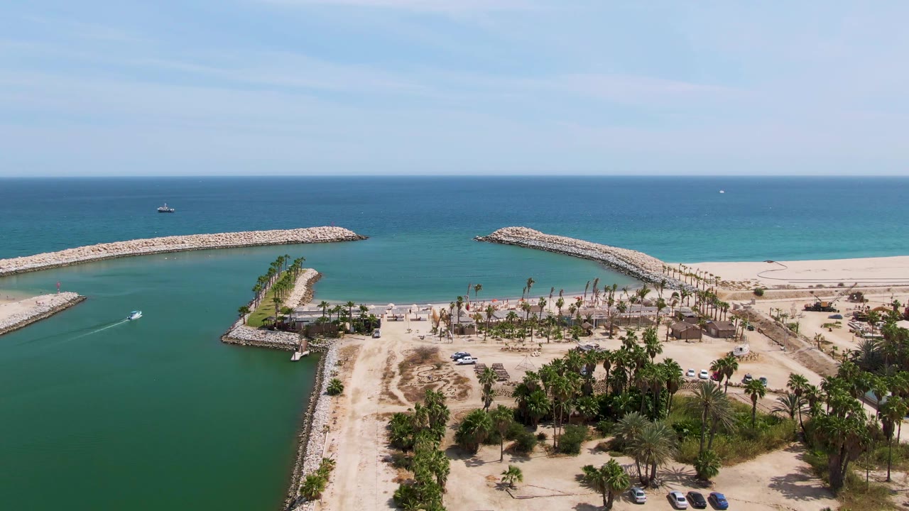 Aerial view over Puerto Los Cabos Beach with rocky breakwaters enclosing turquoise water, a sandy shore, palm trees, and no visible people.
