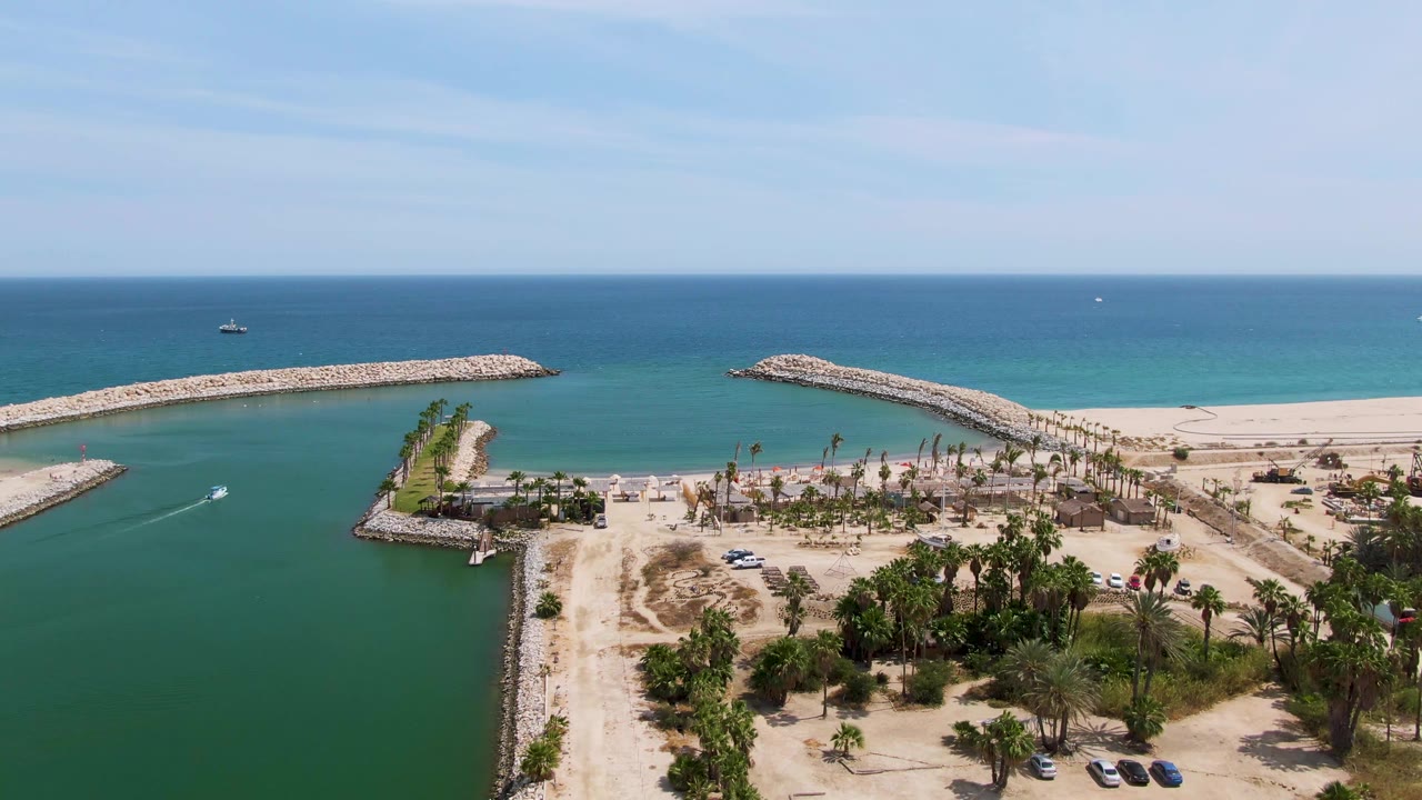 Small boat crosses protected water near rock jetties and a sandy beach.
