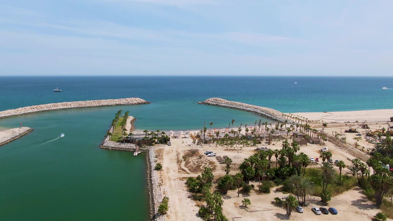 Drone-style aerial frame over Puerto Los Cabos Beach showing a palm-lined sandy shore, turquoise inlet water, long rock breakwaters, distant boats, and no visible people.
