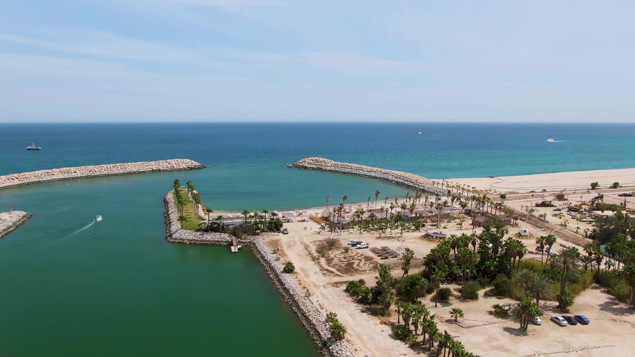 At Puerto Los Cabos Beach in Baja California Sur, a small boat moves through green harbor water beside a rocky breakwater, with sandy shore, palm trees, blue ocean, and no visible people.