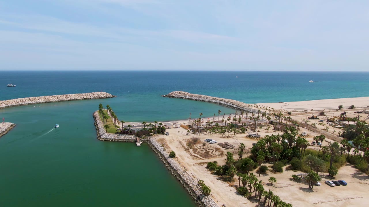 At Puerto Los Cabos Beach in Baja California Sur, the boat sits farther into the harbor channel as its white wake curves across green water, with palm rows, sandy shore, open ocean, and no visible people.