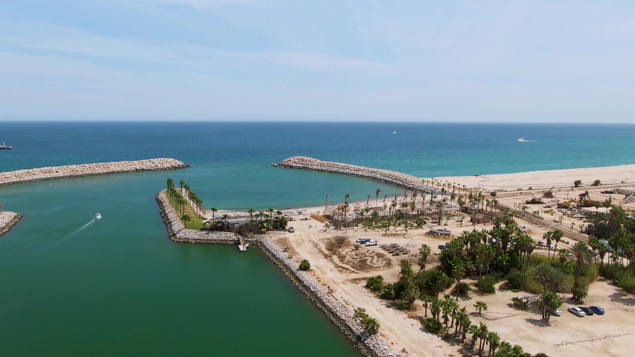 At Puerto Los Cabos Beach in Baja California Sur, a wide aerial frame shows the boat wake inside the green harbor entrance, rocky breakwaters on both sides, sandy beach, palms, deep blue ocean, and no visible people.