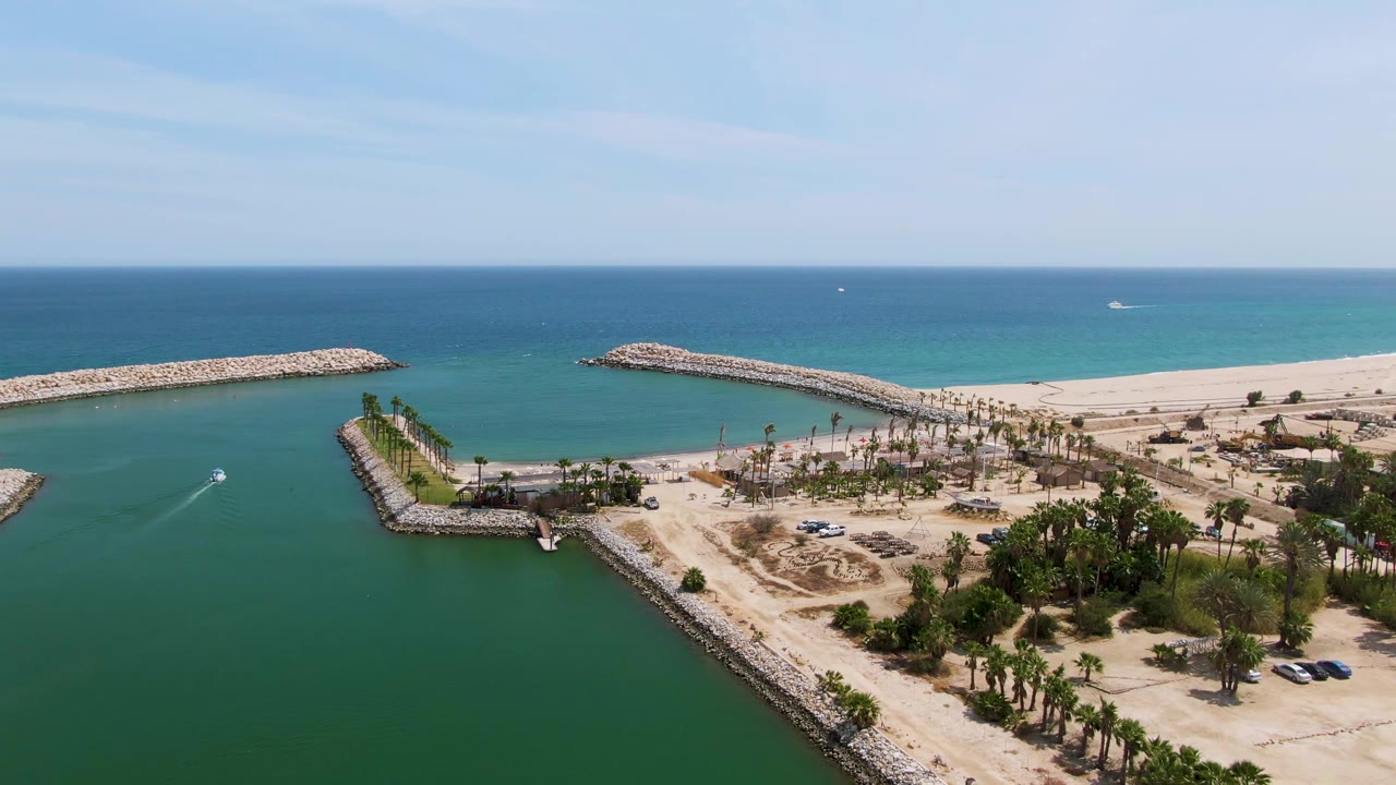 Aerial coastal harbor view with a small boat wake, sandy shoreline, palms, and blue ocean.
