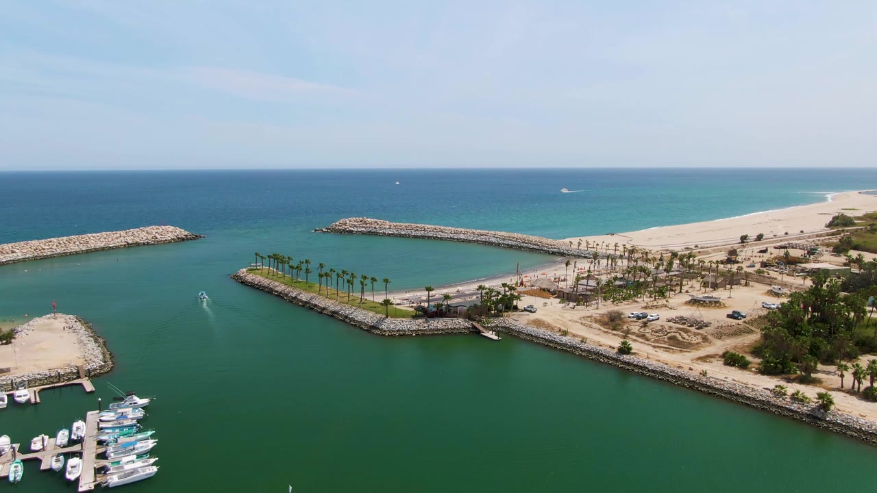 Aerial view at Puerto Los Cabos Beach with a small vessel wake crossing the green channel, docked boats near the lower edge, palms on sand, breakwaters, ocean horizon, and no visible people.