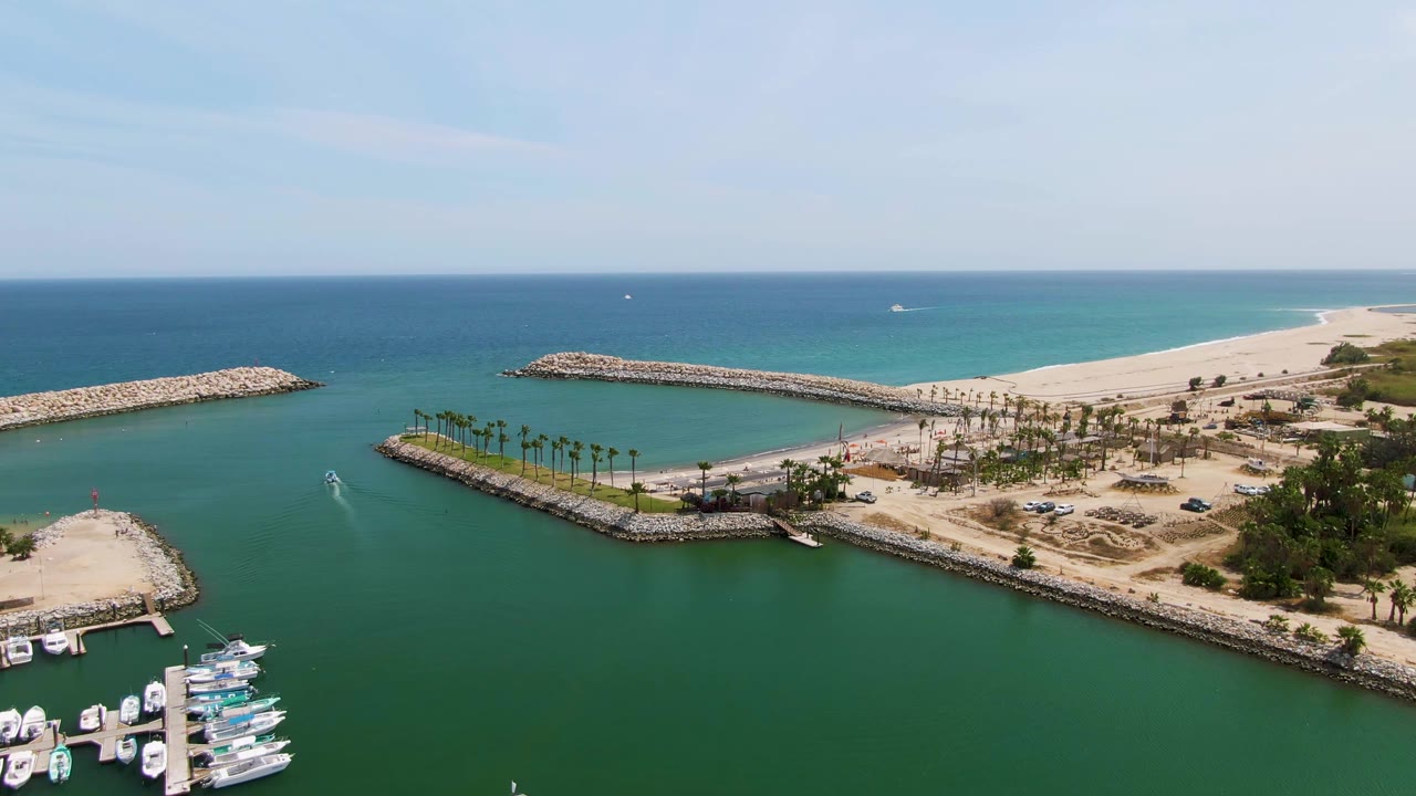 High aerial frame of Puerto Los Cabos Beach with green channel water in front, docked boats at the lower right, a line of palms on sand, two rocky breakwaters, open blue ocean, and no visible people.