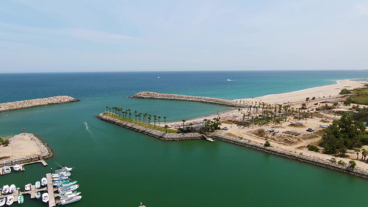 Aerial frame over Puerto Los Cabos Beach with more docked boats visible at lower left, green marina water, a sandy point, palm-lined shore, rocky breakwaters, blue ocean, and no visible people.
