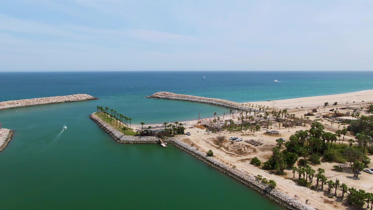 Aerial opening view over Puerto Los Cabos Beach with green harbor water, rock jetties, palm-lined shore, sandy beach, blue ocean, and no visible people.