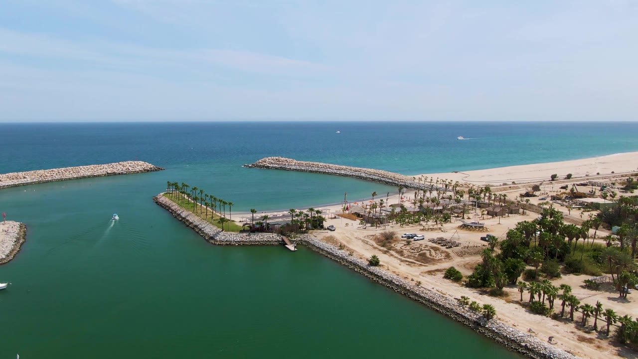 Puerto Los Cabos Beach harbor seen from above as the channel sits between rock breakwaters, with palms and sandy beach beyond, small boats on the water, and no visible people.