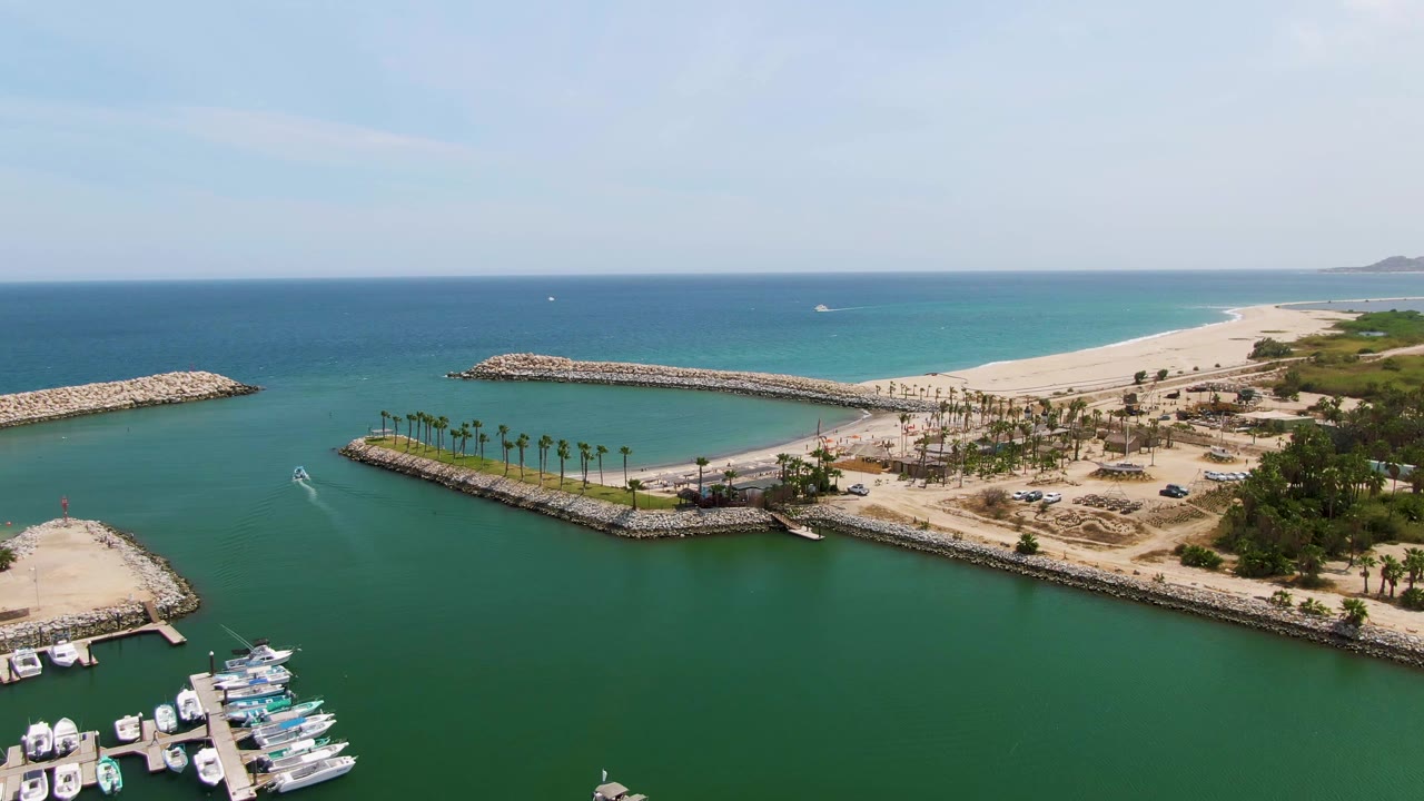 Opening aerial frame over Puerto Los Cabos Beach with docked boats at the lower left, green marina water, rock breakwaters, palm rows, sandy beach, blue ocean, and no visible people.
