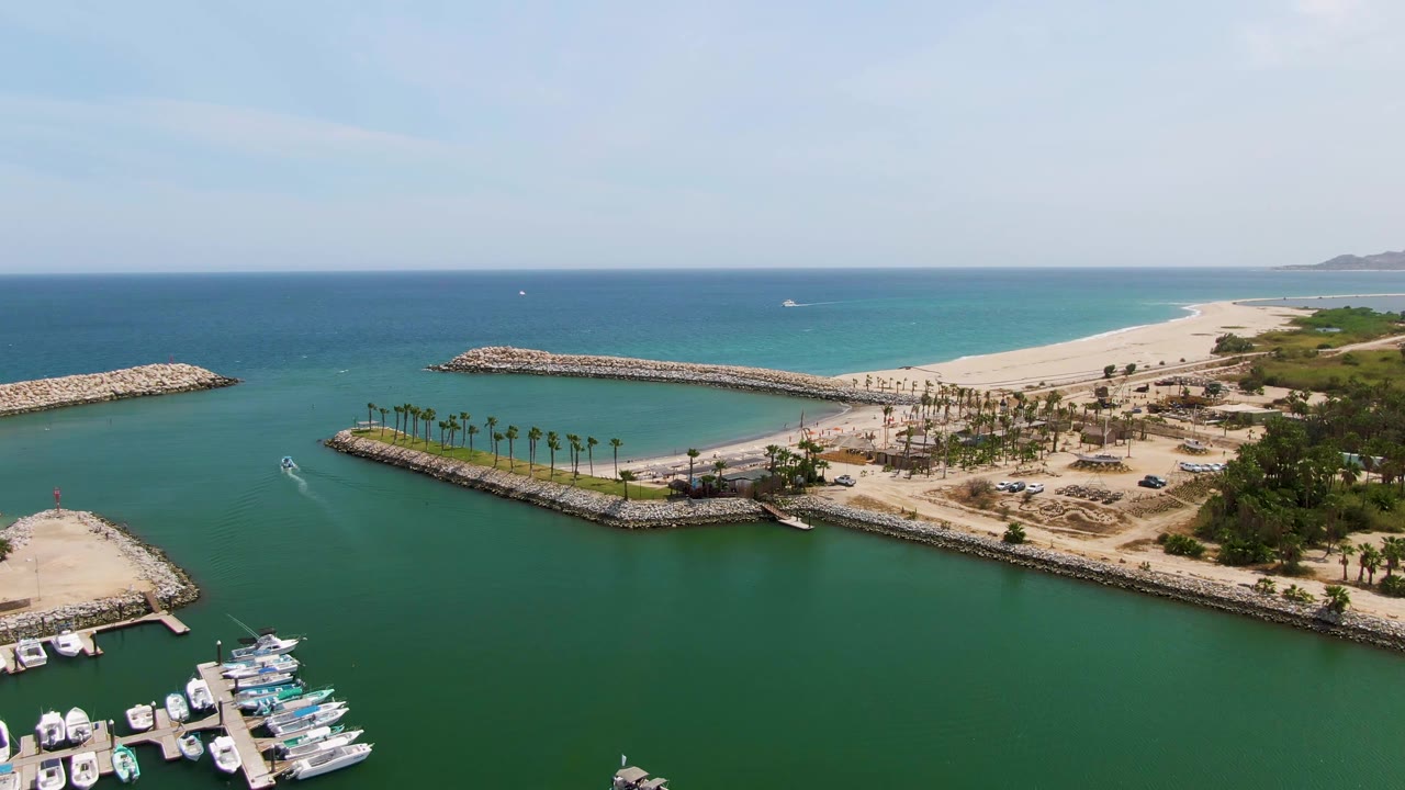 Wide drone view of marina docks, green water, rock jetties, palm-lined shore, and sandy beach.