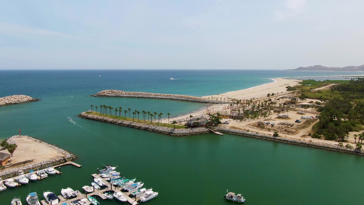 Puerto Los Cabos Beach aerial view with the small boat slightly farther along the channel, marina slips in the lower foreground, palms lining the shore, sandy beach behind the breakwater, and no visible people.