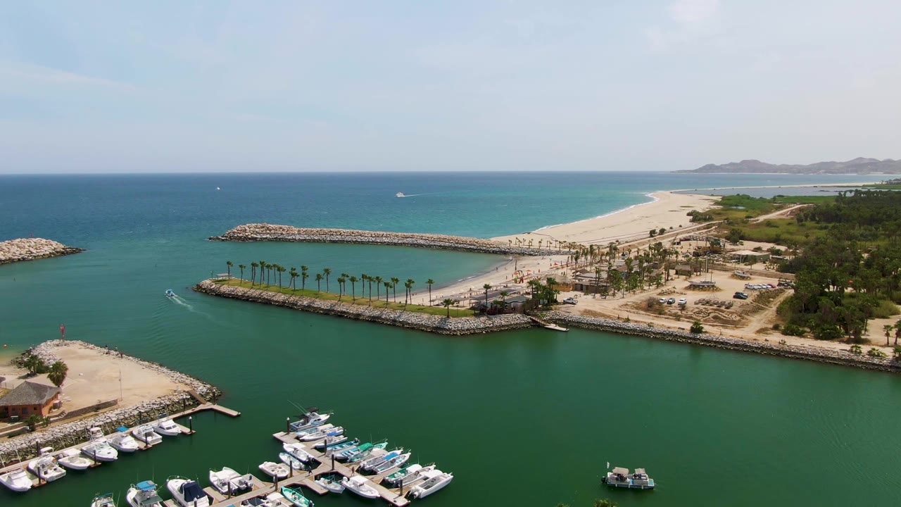 Puerto Los Cabos Beach aerial frame with more marina docks visible in the lower foreground, the small boat crossing the green channel, rocky harbor edges, palms, pale sand, open ocean, and no visible people.