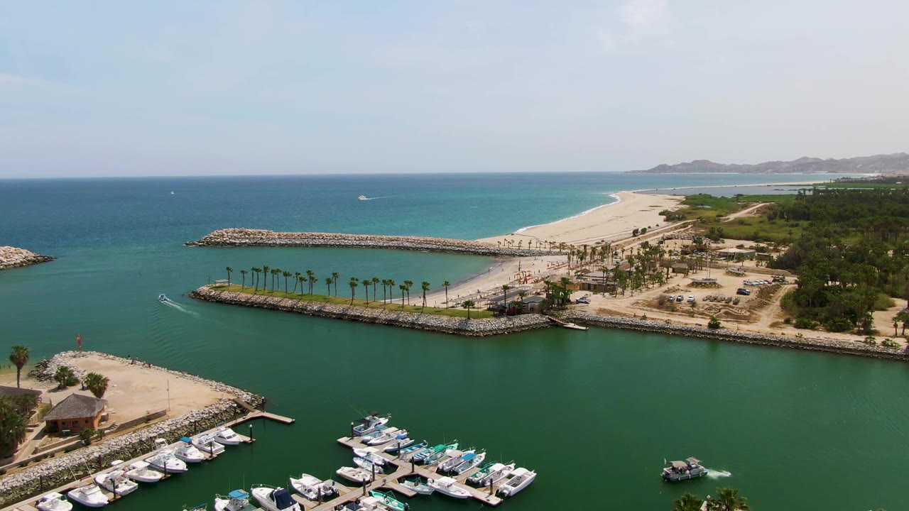 Aerial view of marina boats, turquoise water, breakwater, sandy beach, and ocean horizon.