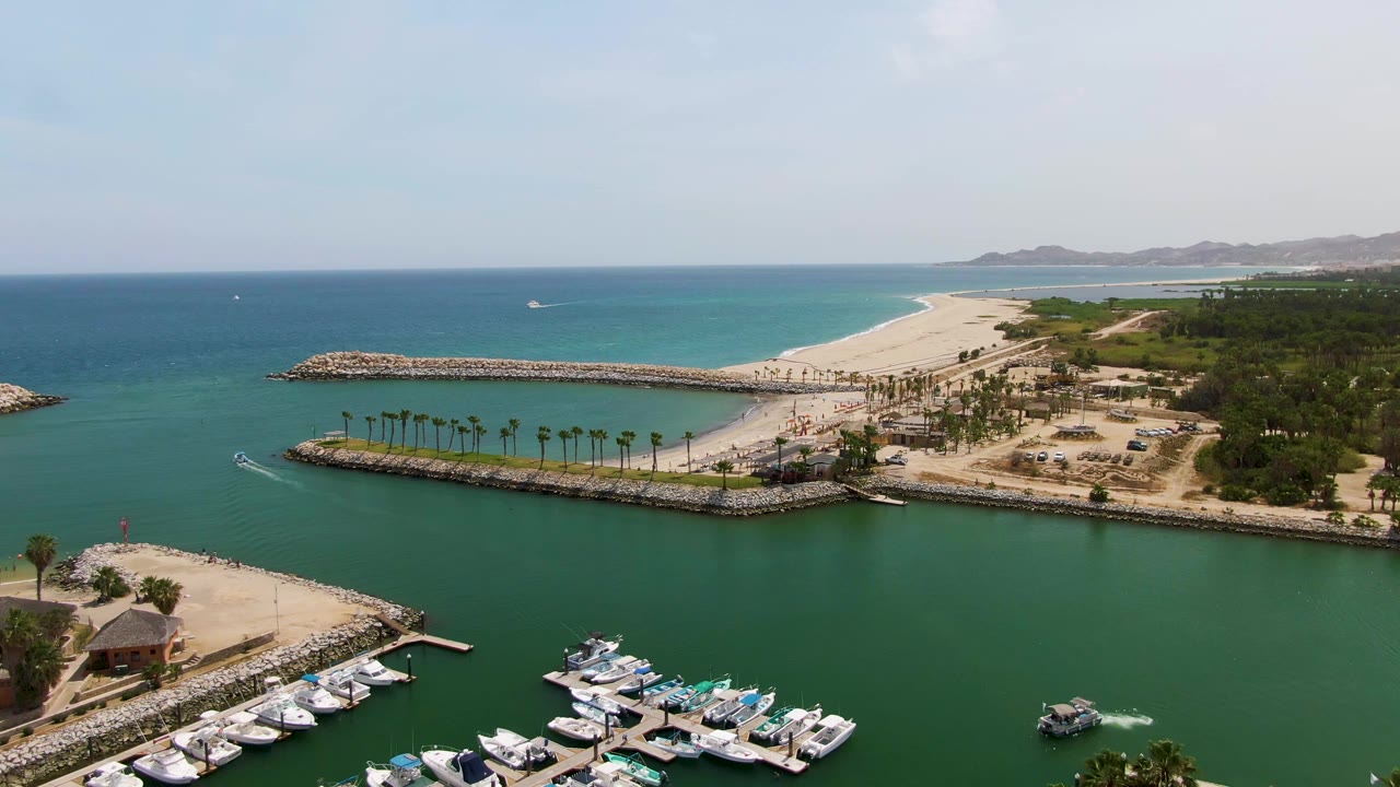 Aerial view over Puerto Los Cabos Beach with marina docks at the bottom edge, a small speedboat wake on the water, palms along the breakwater, sand beyond it, and no visible people.