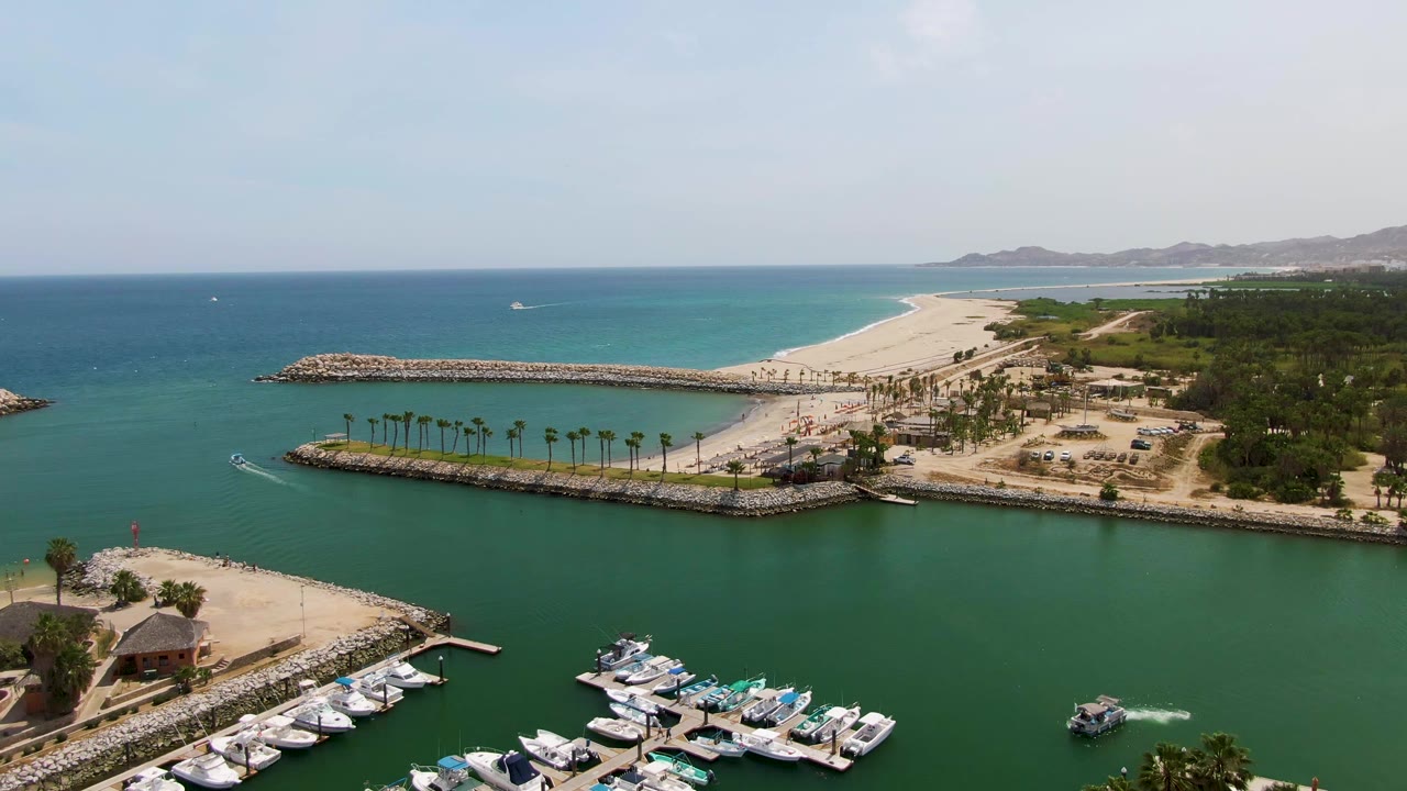 Puerto Los Cabos Beach aerial frame with docked boats clustered at the lower edge, a curved wake near the right side of the water, sandy shoreline, breakwaters, ocean horizon, and no visible people.