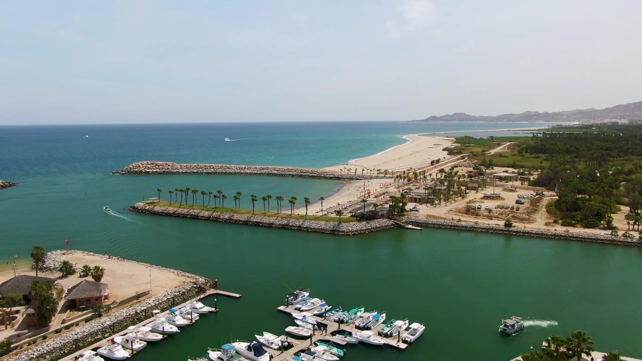 Aerial frame from Puerto Los Cabos Beach showing more open water across the left side, marina boats along the bottom, a long breakwater across the middle, beach and ocean beyond, and no visible people.