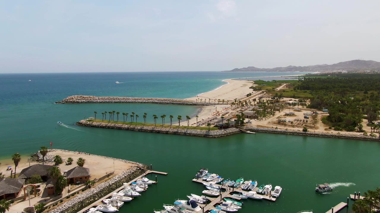 Aerial frame over Puerto Los Cabos Beach, Mexico, showing marina docks, thatched huts, rocky breakwaters, a sandy beach, blue ocean, distant hills, and no visible people.