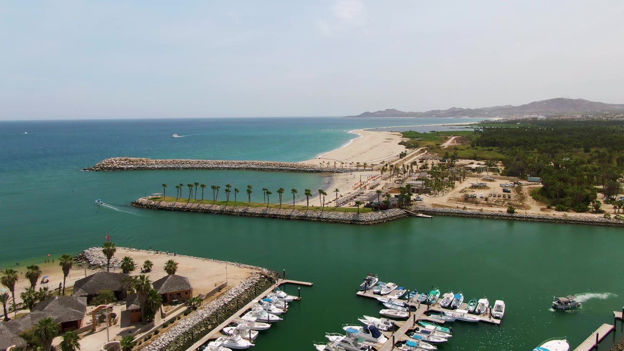 High aerial frame of Puerto Los Cabos Beach showing the marina entrance, parallel breakwaters, moored white boats, turquoise ocean near the sand, low hills, and no visible people.