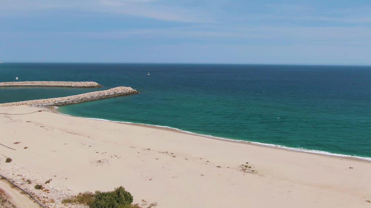 Wide aerial view of Puerto Los Cabos Beach with pale sand in the foreground, two rocky breakwaters offshore, calm blue ocean beyond, and no visible people.