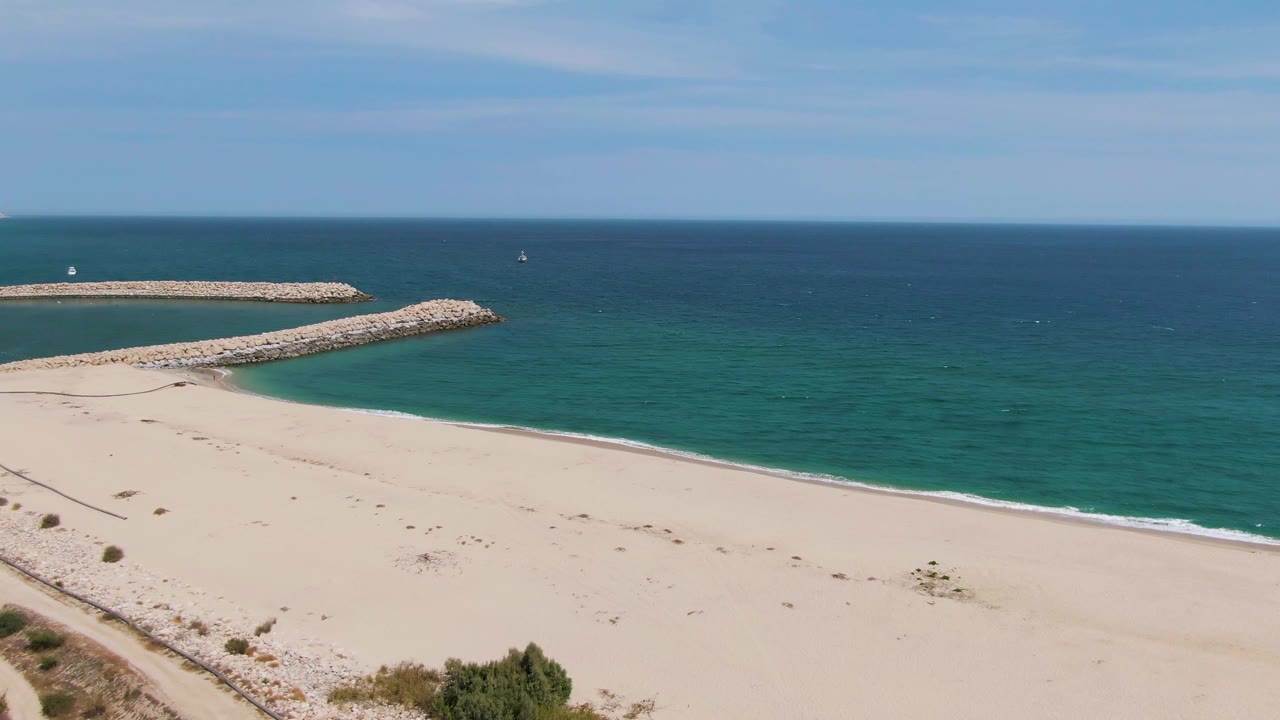 Puerto Los Cabos Beach appears from above with a broader stretch of sand, a curving rocky breakwater, open ocean, sparse dune plants, and no visible people.