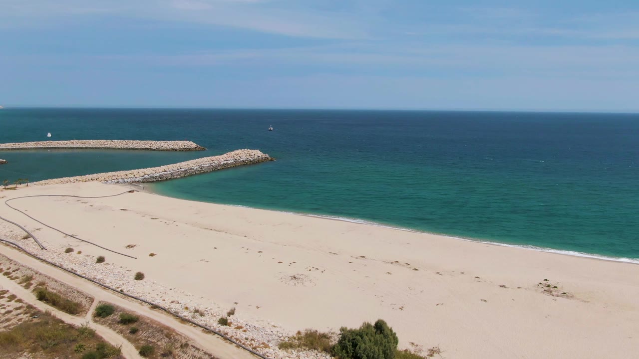 Puerto Los Cabos Beach is seen with the dune path more prominent in the lower frame, pale sand across the center, rocky breakwaters to the left, calm ocean, and no visible people.