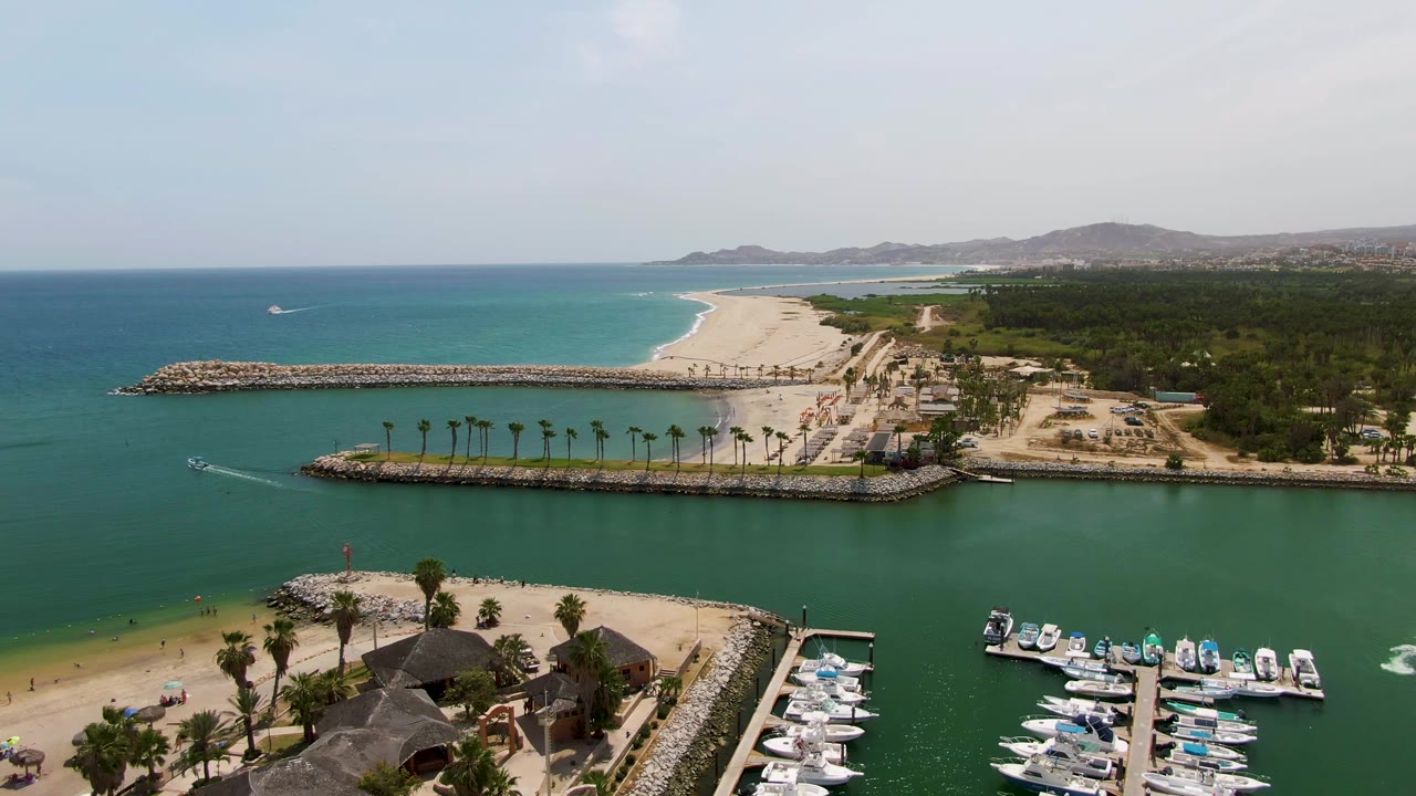 Aerial view over Puerto Los Cabos Beach with marina boats at lower right, sandy beach and umbrellas at lower left, rocky breakwaters, turquoise water, and distant hills.