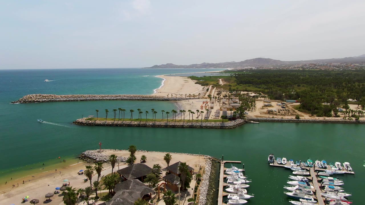 A high aerial frame of Puerto Los Cabos Beach shows a breakwater-lined marina entrance, a row of palms, small beachgoers under umbrellas, docked boats, and bright turquoise water offshore.