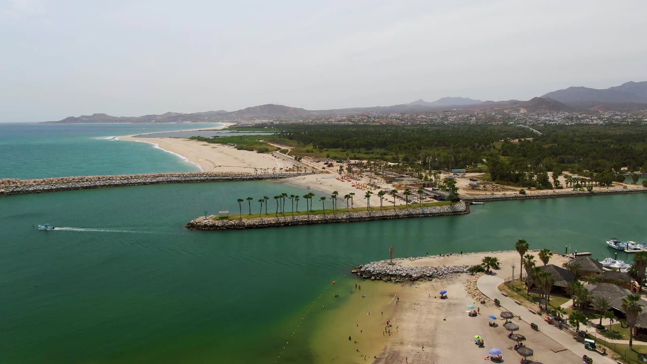 Puerto Los Cabos Beach from above with the boat wake farther across the water, a sandy foreground beach, palm-lined rock breakwater, small umbrellas, parked cars, distant shoreline, and visible people.