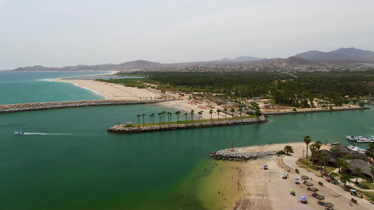 High aerial view of Puerto Los Cabos Beach with curved turquoise shoreline at left, green protected water below, breakwaters through the center, boats near the right edge, palm trees, parked cars, and visible beachgoers.