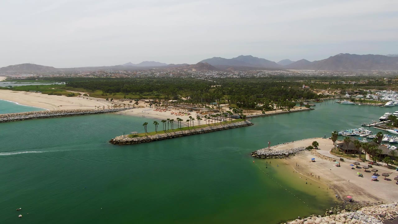 Aerial frame at Puerto Los Cabos Beach showing a bright boat wake stretching across green lagoon water, sandy beach areas on both sides, marina docks to the right, palms, and distant mountains with no visible people.