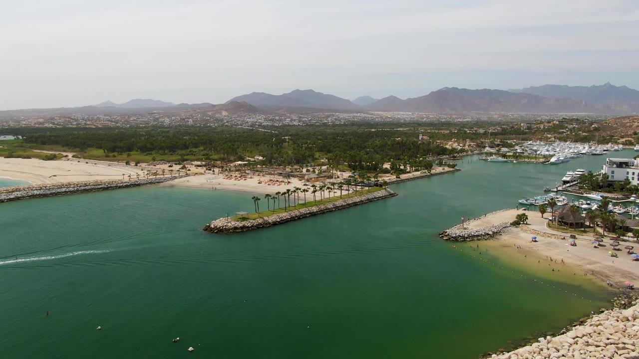 Opening aerial frame over Puerto Los Cabos Beach with green marina water in the foreground, a sandy shore and rocky breakwater at midframe, boats near a white waterfront building, distant mountains, and no visible people.