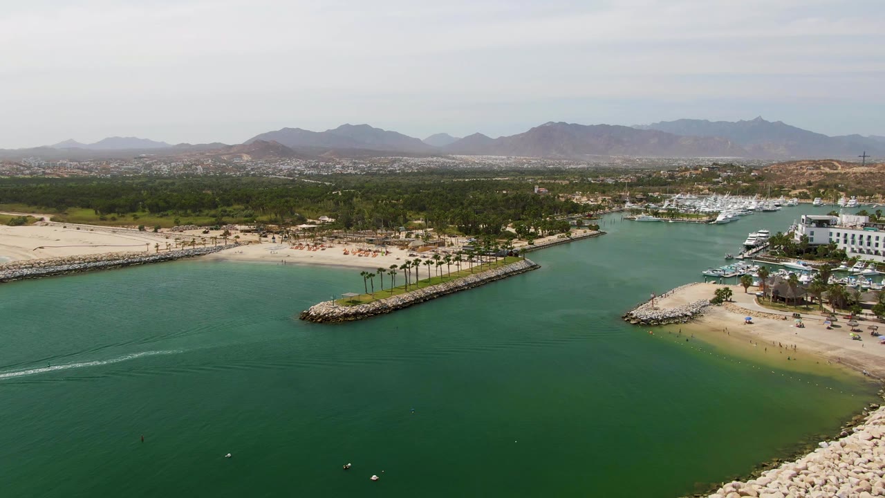 At 0:02, the aerial view holds over Puerto Los Cabos Beach as faint wake lines cross the green water, with a sandy shoreline, breakwaters, marina boats, palms, a white building, mountain ridges, and no visible people.