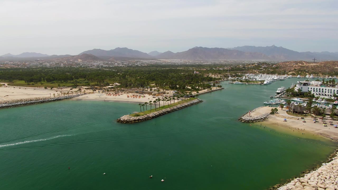 Aerial harbor scene with green water foreground, beach and breakwaters, marina boats, palms, and mountains.