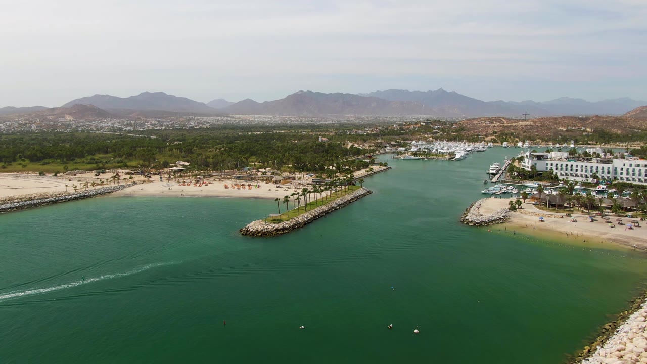 Aerial frame over Puerto Los Cabos Beach showing turquoise water, a white boat wake entering from the left, sandy shoreline, rocky breakwater, marina buildings, palm trees, distant mountains, and no visible people.