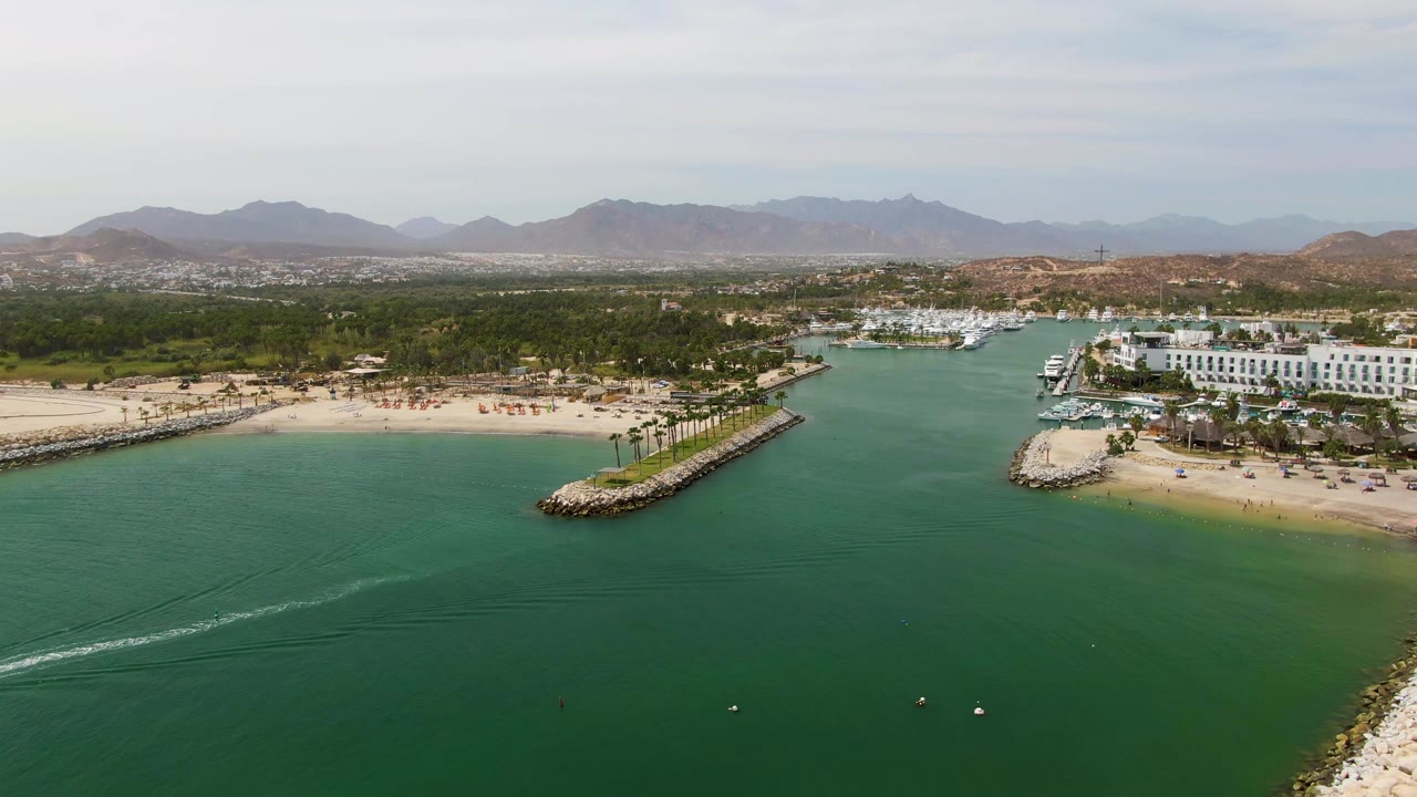 Puerto Los Cabos Beach aerial view with the white wake extending farther across green-blue marina water, sandy beach and breakwater at midframe, marina structures to the right, mountains beyond, and no visible people.