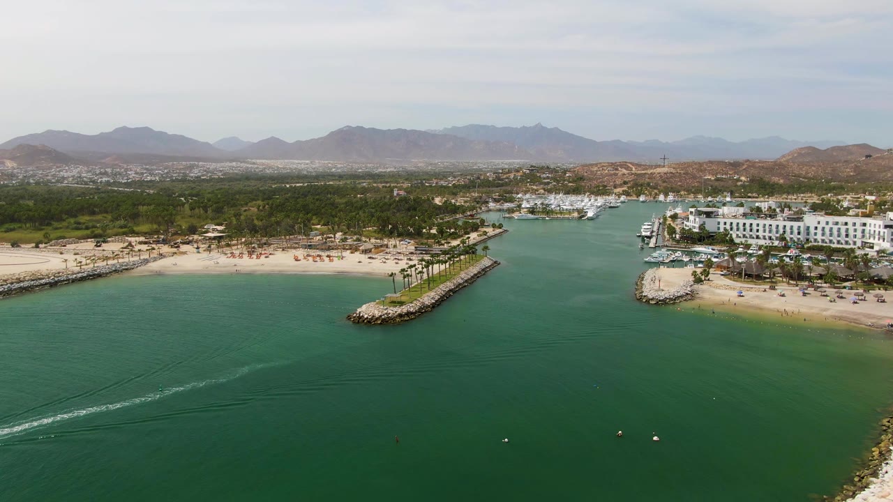 Wide aerial frame at Puerto Los Cabos Beach showing the wake cutting diagonally through turquoise water, a curved sandy shore, a rocky breakwater point, white marina buildings, palms, mountains, and no visible people.