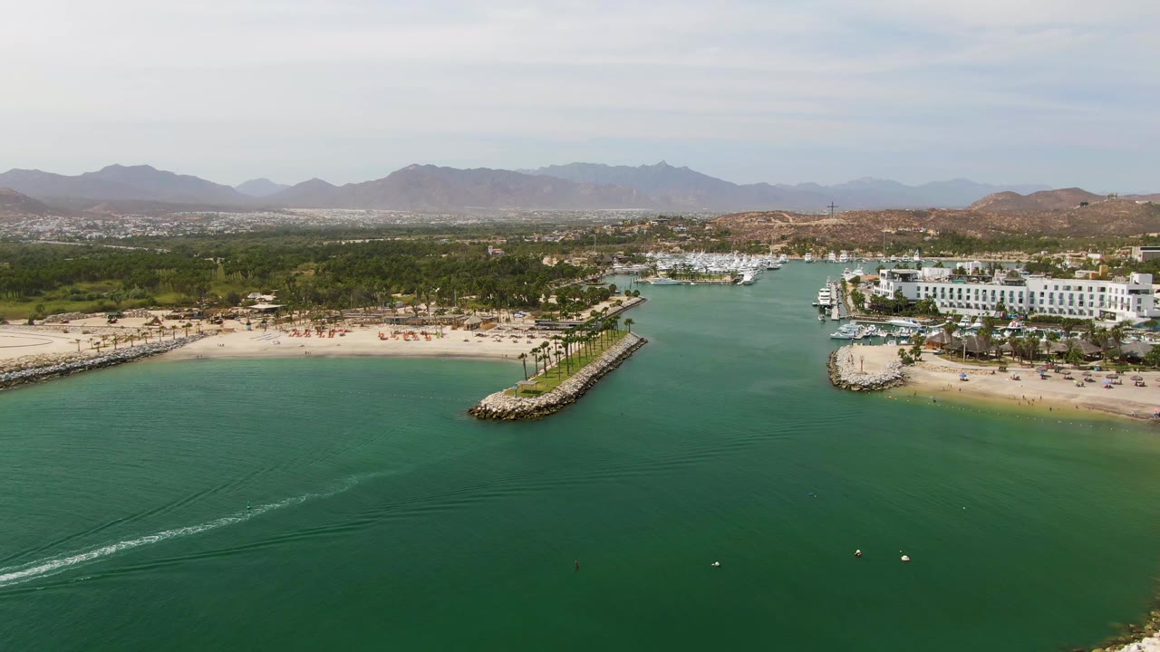 Puerto Los Cabos Beach frame with the boat wake trailing across the lower left water, sandy beach and palms along the center, marina buildings to the right, hazy mountain ridges behind, and no visible people.